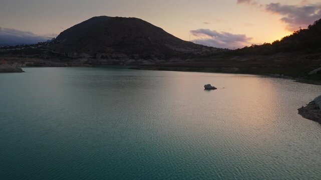 Drone video of a tranquil mountain lake in Spain during sunset, with still turquoise water, rocky shoreline, and soft evening light reflecting on the surface.