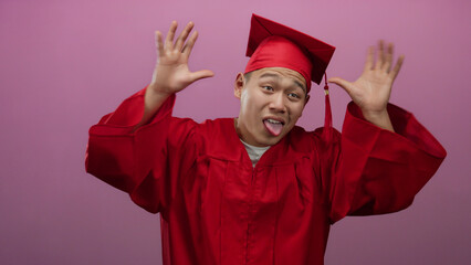 Young man in red graduation gown making playful faces against pink wall, celebrating graduation day.
