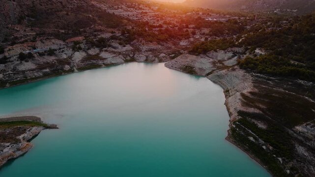 Drone video of a tranquil mountain lake in Spain during sunset, with still turquoise water, rocky shoreline, and soft evening light reflecting on the surface.