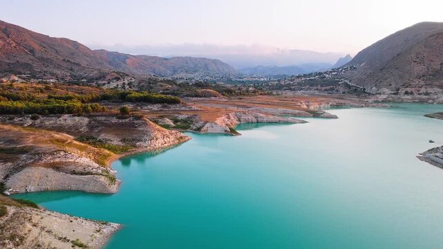 Drone video showing a vivid turquoise lake in Spain with rocky, sunlit shoreline and earthy cliffs creating striking contrast between water and land.