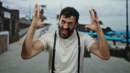 Hispanic man with a beard gesturing emotionally on a seaside promenade with a beach and sea in the...