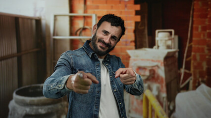 Hispanic man with beard smiling and pointing in a friendly manner at a construction site wearing a denim jacket with brick and tools in the background creating a casual and engaging scene.