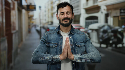 Hispanic man with beard stands thoughtfully on urban street, wearing denim, expressing emotion...