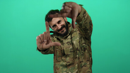 Hispanic man with beard in military uniform poses with green background creating a frame with his...