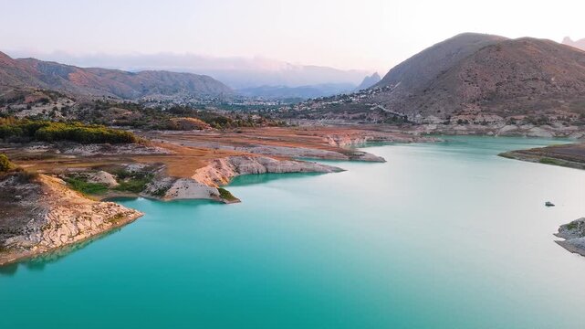 Drone video showing a vivid turquoise lake in Spain with rocky, sunlit shoreline and earthy cliffs creating striking contrast between water and land.