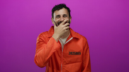 Hispanic man in an orange prison jumpsuit with a beard stands against a vibrant pink background, exuding a juxtaposition of confinement and light-heartedness while covering his smile.