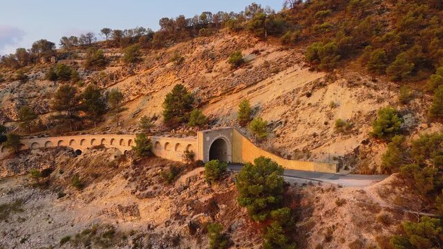 Drone video of a historic stone tunnel and arched wall along a mountain road in Spain, lit by warm golden hour sunlight.