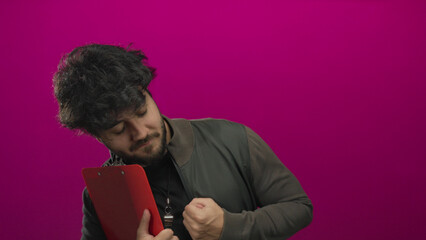 Young hispanic man with beard holding clipboard against pink background wearing jacket and whistle...