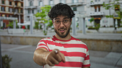 Young man with beard smiling outdoors on a street wearing glasses in a red striped shirt pointing happily on a bright day embracing fun and confidence with vibrant ambiance.