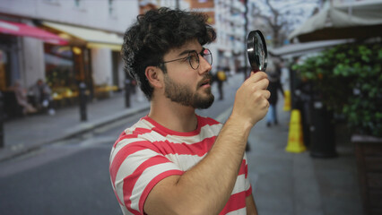 Young man with beard examines street scene using magnifying glass, wearing striped shirt outdoors,...
