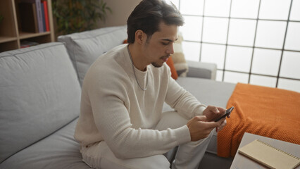 Young man indoors using smartphone sitting on a couch in a modern living room with a notebook on...