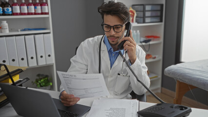 Hispanic man in clinic wearing lab coat talks on phone while reviewing invoices at desk with laptop...