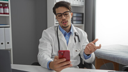 Young hispanic doctor man in clinic using smartphone for telemedicine consultation indoors with focused expression showing modern medical technology in professional setting