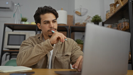 Young man working with laptop in an office, appearing thoughtful and focused, surrounded by various...