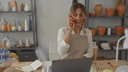 Young woman holding phone to ear in pottery studio while checking orders and reaching for clay...