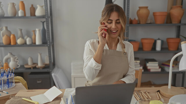 Young woman holding phone to ear in pottery studio while checking orders and reaching for clay tools; creativity focus serenity. - Powered by Adobe