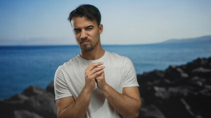 Young man at the seaside with contemplative expression, wearing casual white shirt against a serene...