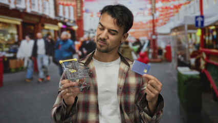 Young man outdoors holding a credit card and toy shopping cart on a busy city street highlighting urban shopping context in vibrant market ambiance.