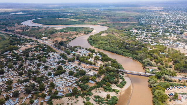 Aerial view of the brown Tana River snaking through the arid landscape, contrasting with the dense green vegetation and buildings, Garissa, Kenya.