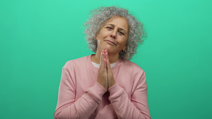 Grey-haired middle-aged woman making a pleading gesture with clasped hands against an isolated green wall background.
