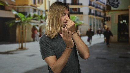 Blond man with long hair pinches nose and waves free hand in disgust on a bustling street; stinky...