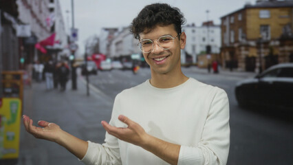 Young hispanic man wearing glasses and white sweater smiling with hand showing thumbs up on city...