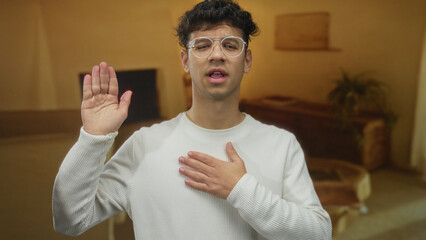 Young man in white shirt and glasses raises hand and touches chest in a cozy residence inside a...