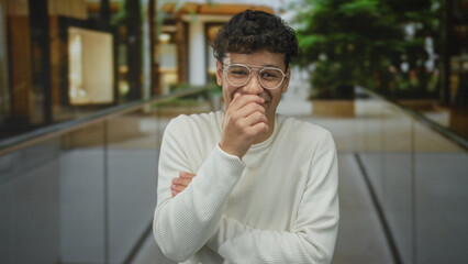 Young hispanic man in white sweater and glasses pinches nose with folded arms on busy city street walkway by storefront windows; disgust.