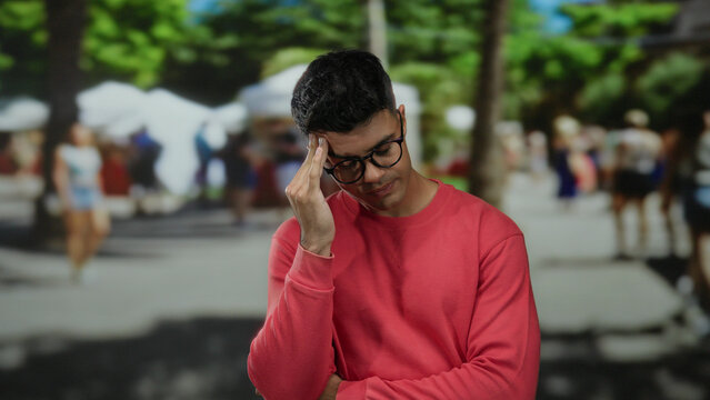 Young man in neon shirt stands thoughtfully in busy city street surrounded by people and trees.
