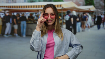 Fototapeta premium Woman in pink sunglasses poses confidently at an outdoor market, capturing a lively street scene with people in the background, emphasizing a vibrant shopping atmosphere.