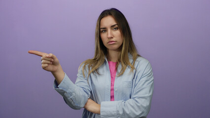 Young blonde woman points assertively against an isolated purple wall in a confident expression.