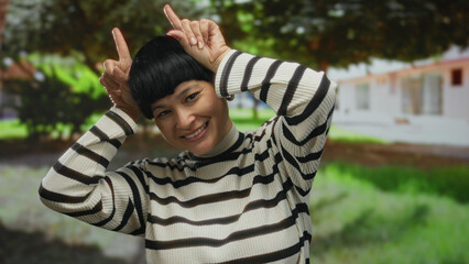 Woman smiling in an outdoor park setting with greenery, playfully posing with hands making bunny ears in vibrant daylight.