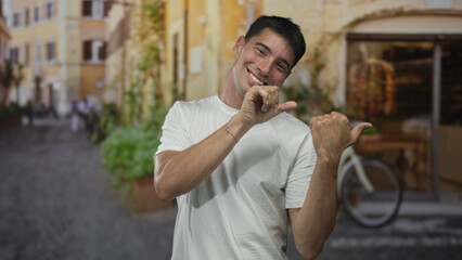 Man wearing white tshirt pointing thumb to side on street with old town building; happiness travel...