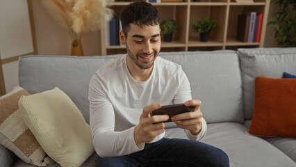 Young man happily playing on a smartphone in a cozy modern living room, surrounded by decorative pillows and plants, showcasing a relaxed indoor atmosphere.