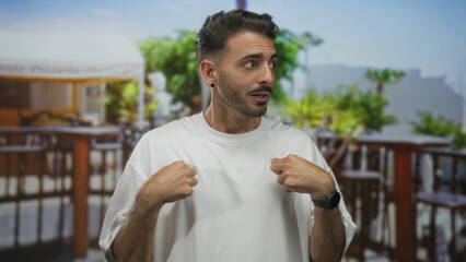 Man in white t shirt points hands to chest and raises eyebrows on a sunny terrace by wooden...