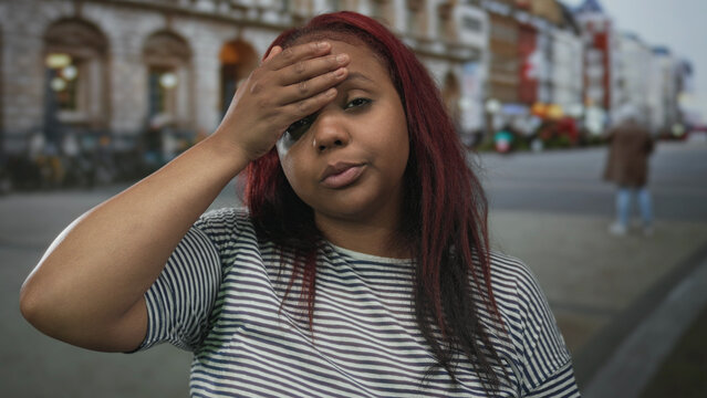 Young african american woman with red hair wearing striped shirt touches forehead and shrugs hand in busy city street; annoyance.