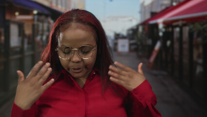 Fototapeta premium Woman wearing red shirt and round glasses fans bare face with hands on restaurant terrace walkway outdoors; heat discomfort.