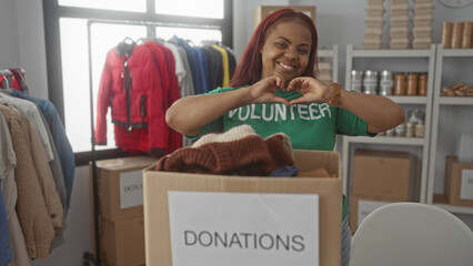 Woman volunteer in green shirt makes heart gesture with hands over cardboard donation box inside bright building; compassion.