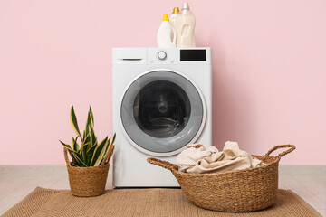 Modern washing machine with houseplant, basket and laundry near pink wall