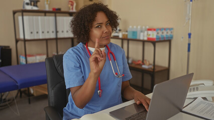 Woman doctor wearing blue scrubs and red stethoscope typing on laptop at desk in clinic; compassion...