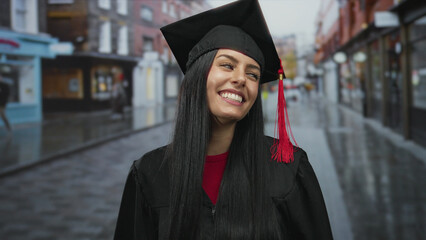 Hispanic woman in graduation cap smiles on city street celebrating achievement in vibrant urban...