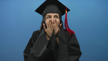 Young hispanic woman in graduation cap and gown celebrating against an isolated blue background,...