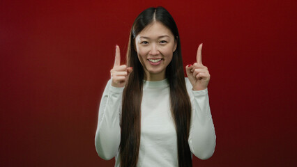 Woman pointing upward on a vibrant red wall background, expressing positive gesture with a smile,...
