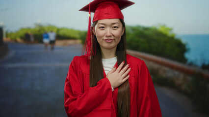 Woman in red graduation gown standing on seaside promenade with long hair and background foliage.