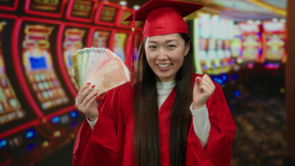 Woman in red graduation gown holding philippine banknotes in a casino with slot machines in the background, smiling and celebrating indoors.