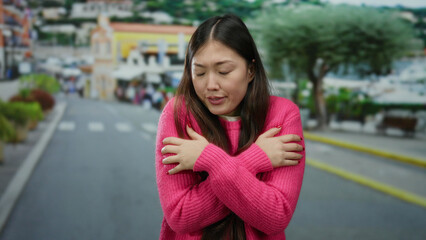 Fototapeta premium Woman in pink sweater in urban street setting with arms crossed appearing cold with blurred cityscape background.