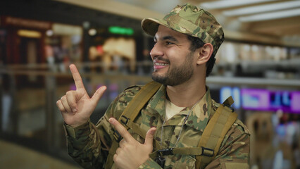 Man in camouflage uniform points finger to side and smiles in shopping center corridor indoors;...