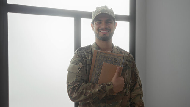 Man in camouflage uniform holds stack of books with one hand against window frame inside building; pride duty. - Powered by Adobe