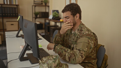 Young hispanic soldier covering mouth yawns at cluttered office desk inside room lined with...