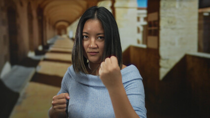 Woman practicing martial arts in an old university hallway, expressing determination, with archways in the background, showcasing strength and focus indoors.